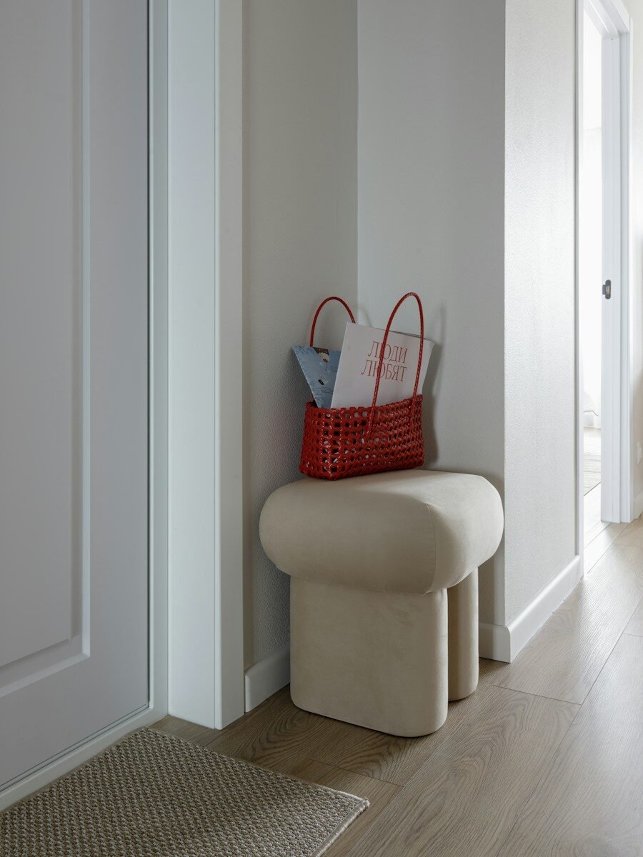 A red woven basket with magazines on a beige upholstered stool in a hallway